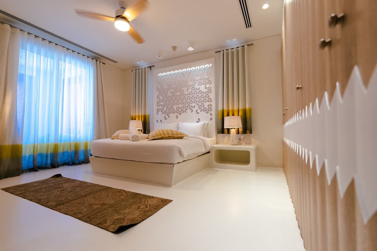 A modern bedroom with a bed, bedside lamps, a ceiling fan, and a decorative wall panel.