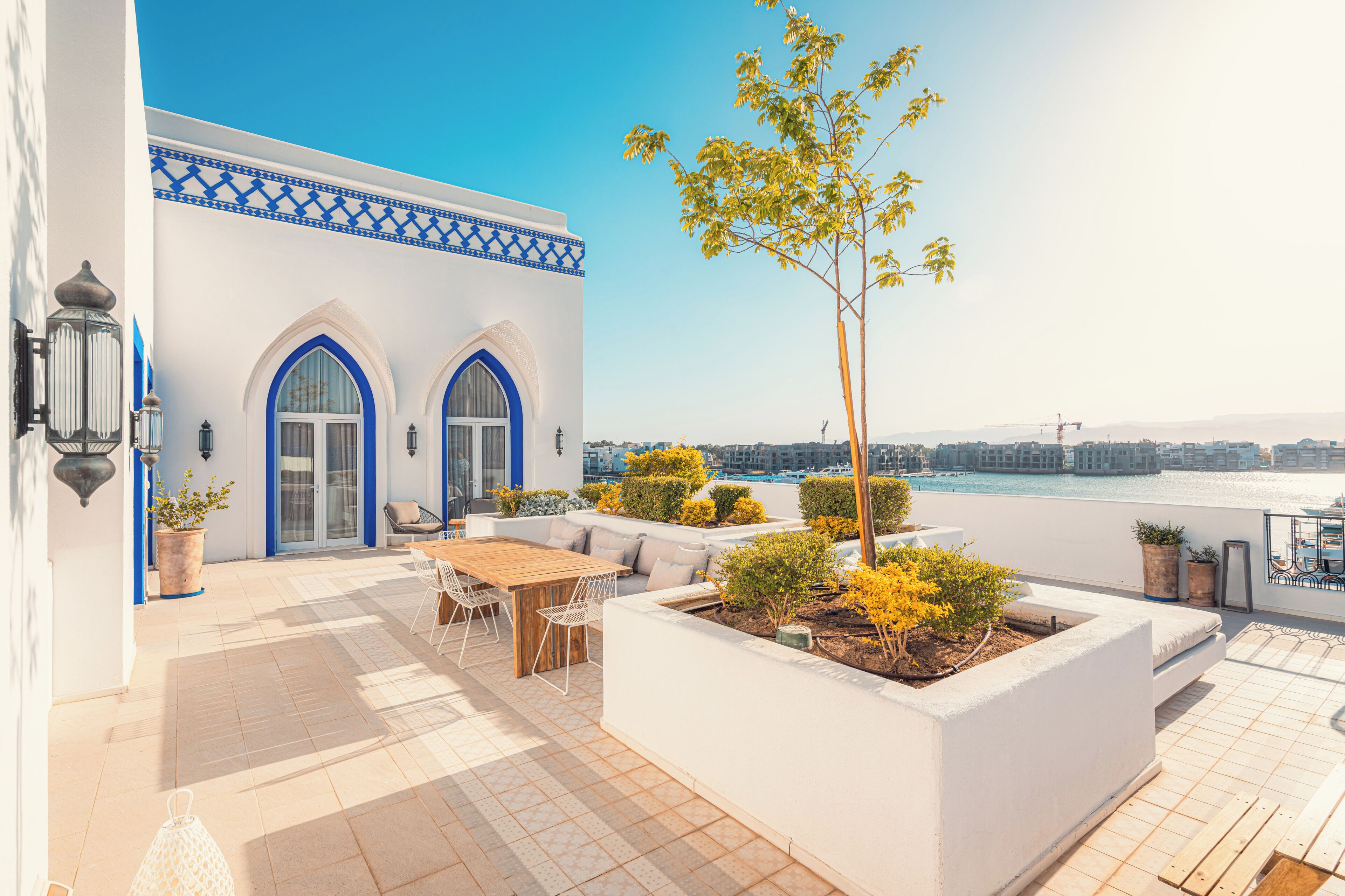 A rooftop terrace with a wooden table and chairs, a white planter with plants, and a view of the sea.