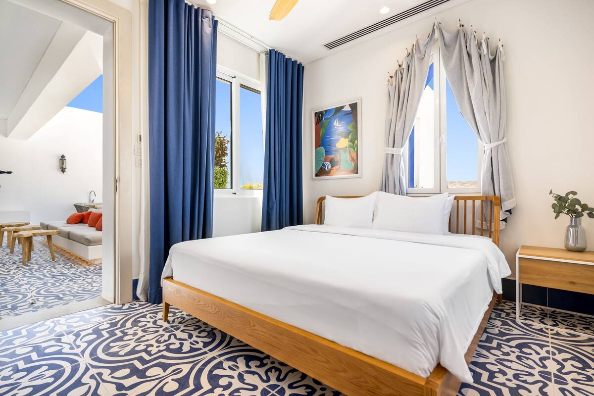 A bedroom with a bed, a bedside table, a window with curtains, a painting on the wall, and a view of the outdoors.