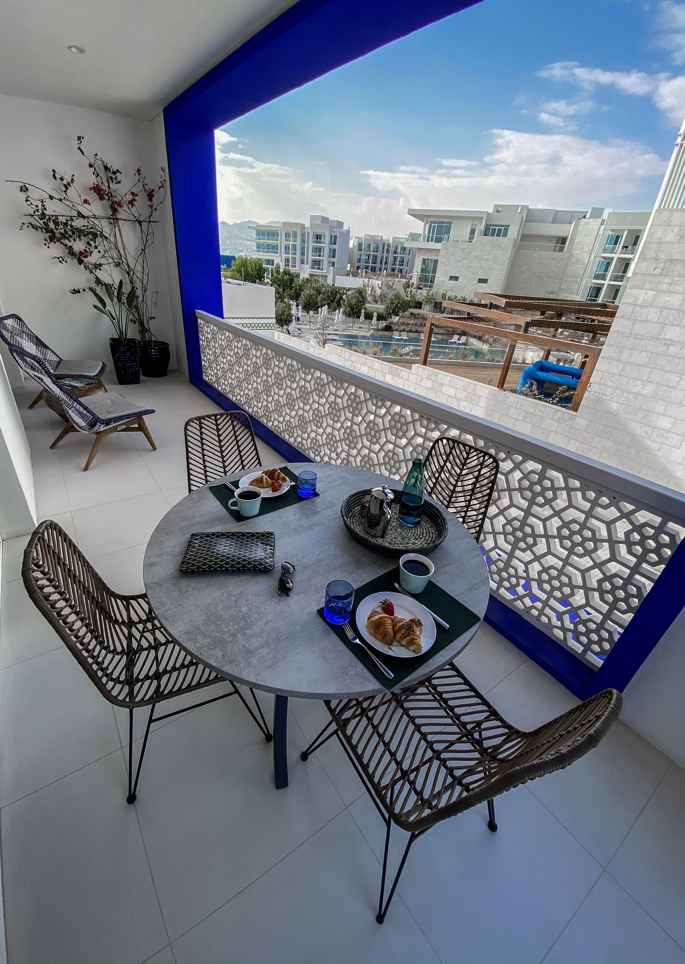 Un balcon avec une table à manger pour deux, entouré de chaises en osier. On y trouve une plante en pot et, en arrière-plan, on aperçoit une piscine et des bâtiments.