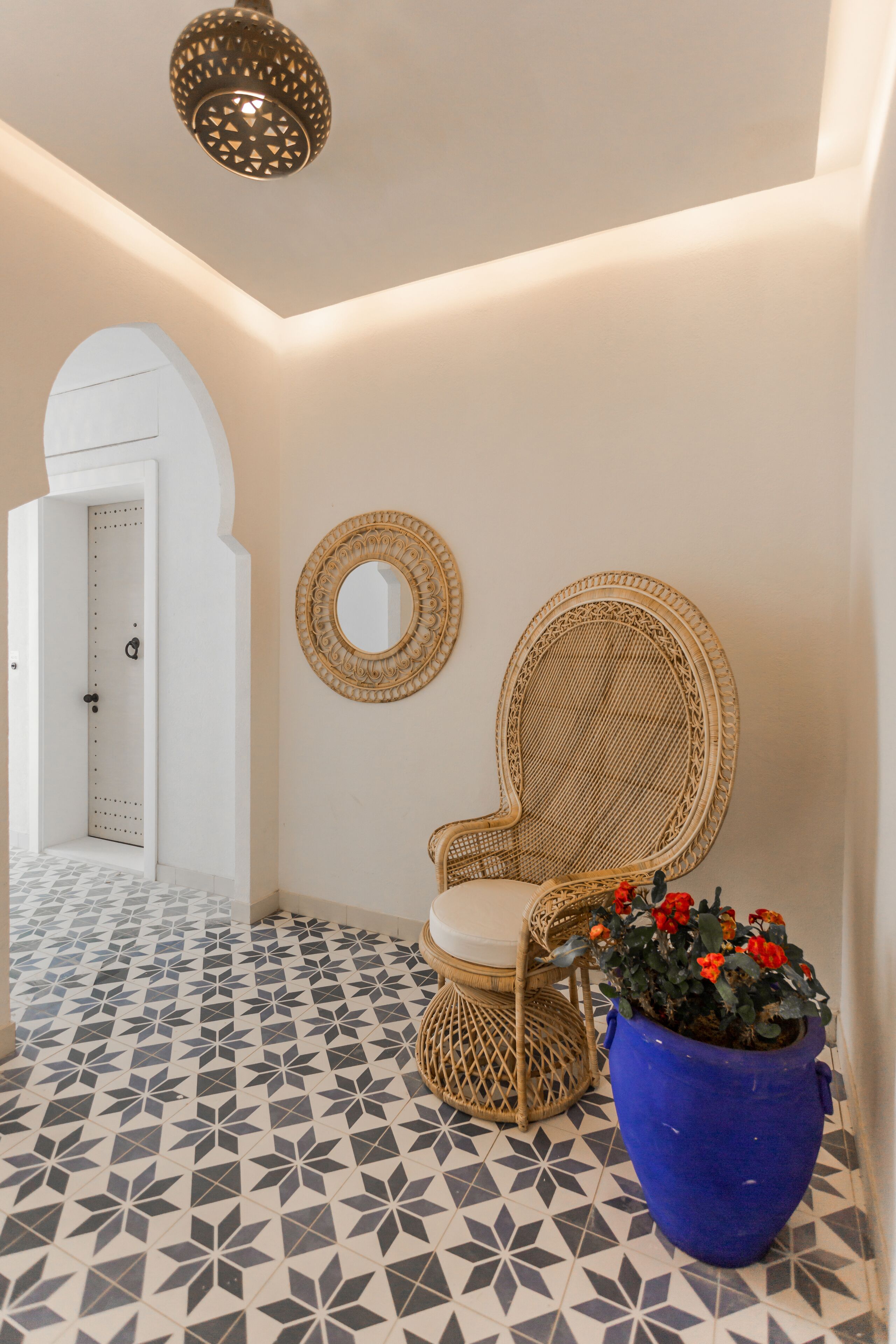 Un pasillo con suelo de diseño geométrico, una silla de mimbre, una planta azul en maceta y un espejo adosado a la pared.