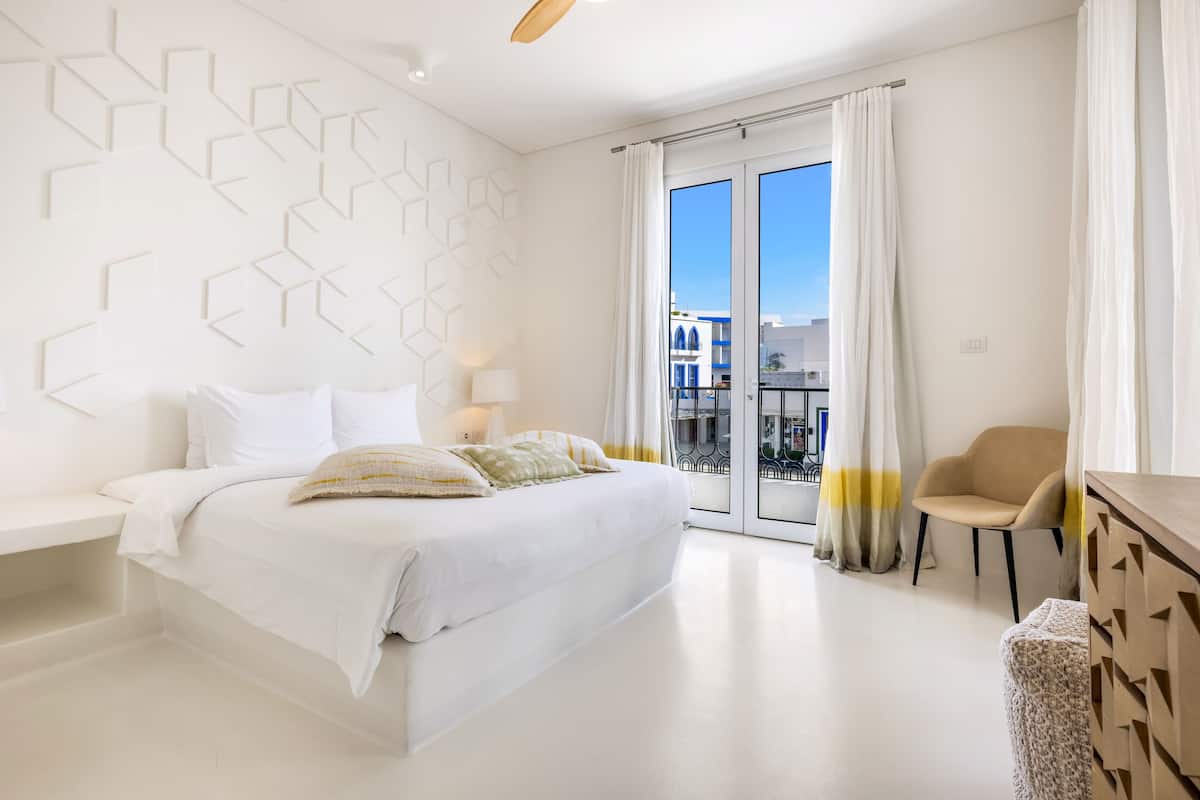 A modern hotel room with a bed, a desk, a chair, and a view of the city through large windows.
