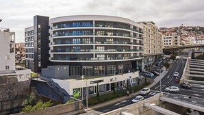 A modern multi-story building with balconies, located near a busy road with vehicles and a pedestrian crossing. - In the Funchal city center, Santa Luzia I (Funchal)