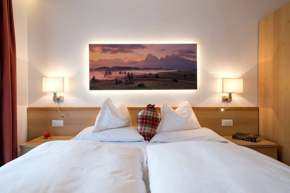 A hotel room with a large bed, two bedside lamps, a painting of a landscape, and a wooden headboard.