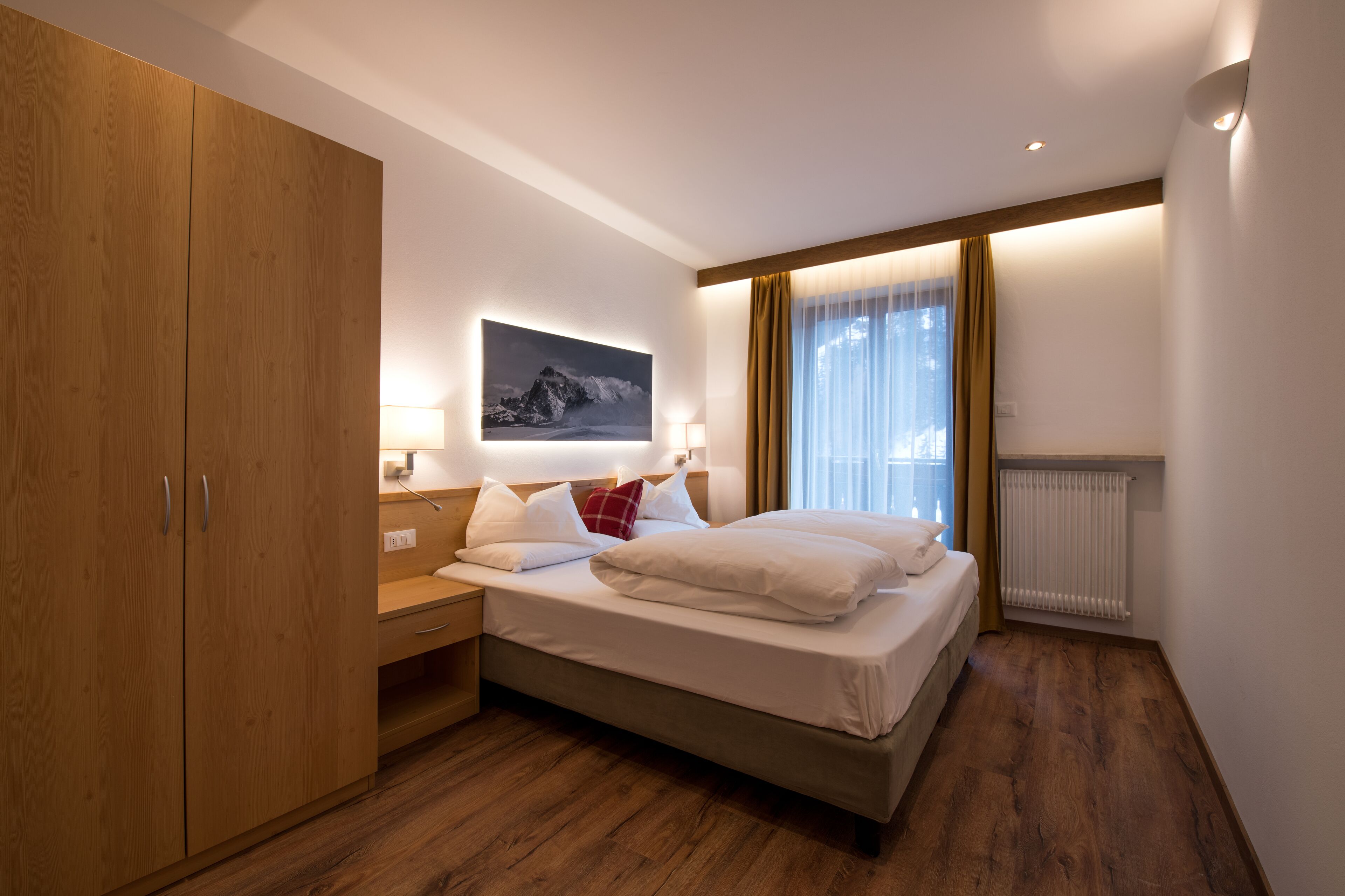 Ein Schlafzimmer mit einem Holfischschrank, einem Bett mit weißer Bettwäsche, einem Nachttisch, einer Wandlampe und einem Fenster mit Vorhängen.