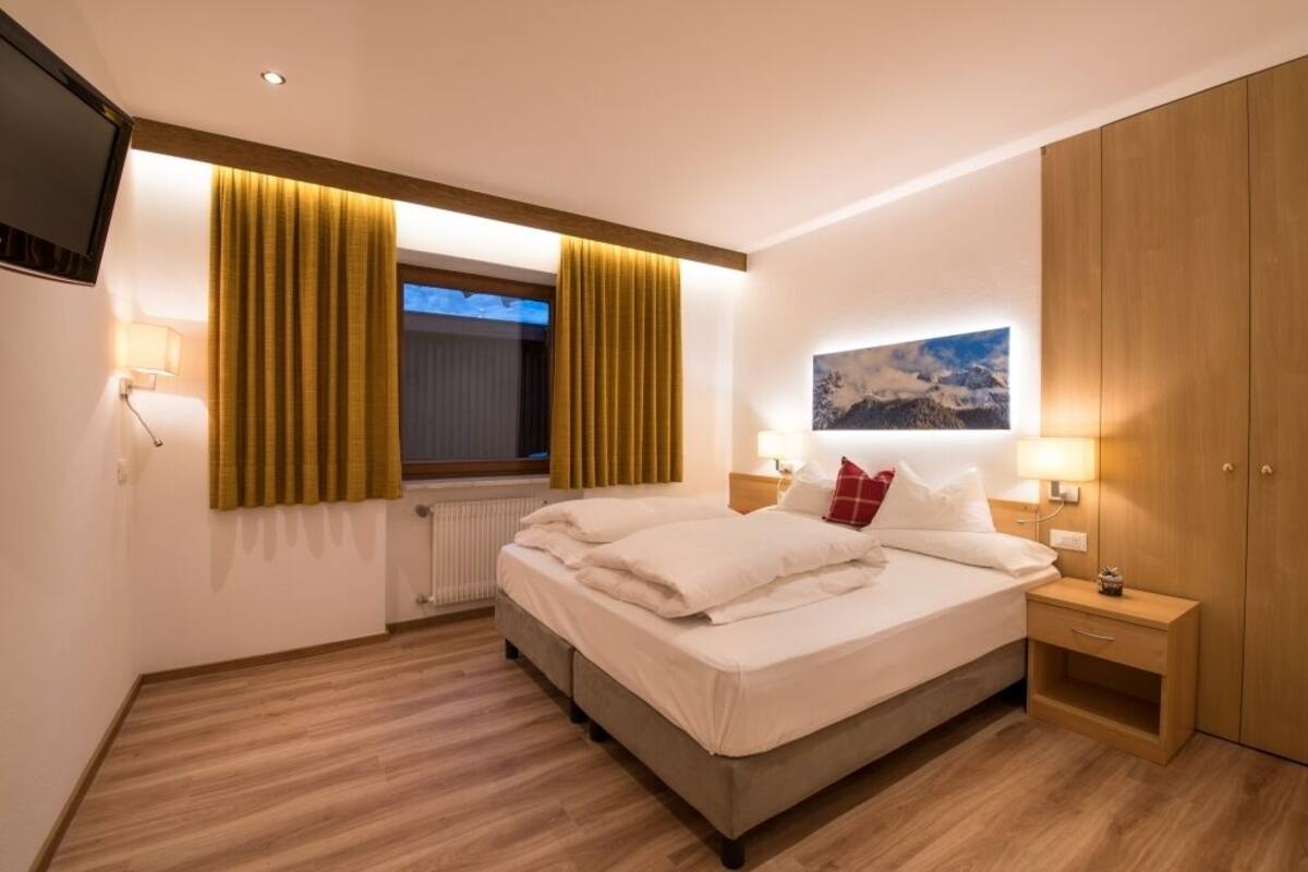 A modern hotel room with a large bed, wooden flooring, a window with curtains, a mounted TV, and a painting on the wall.