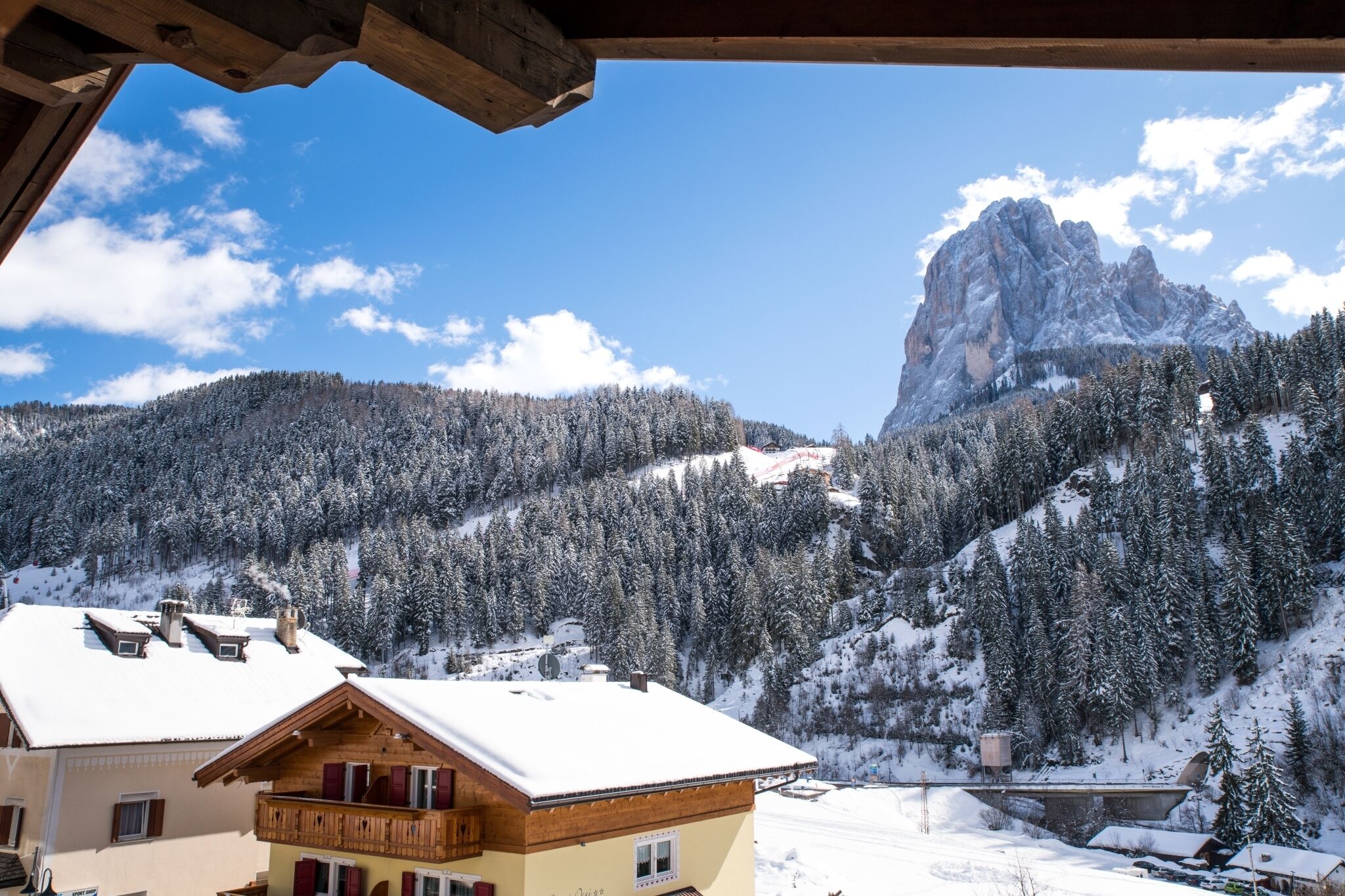 Una cabaña de montaña con balcón de madera y tejado cubierto de nieve, con vistas a un valle boscoso y un imponente pico montañoso.
