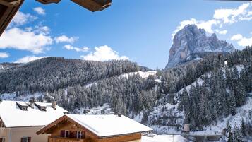 Una cabaña de montaña con balcón de madera y tejado cubierto de nieve, con vistas a un valle boscoso y un imponente pico montañoso.