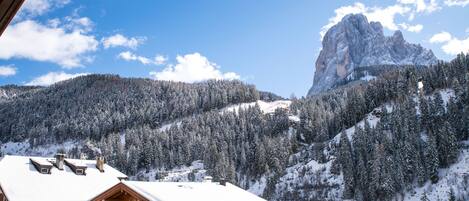 A mountain cabin with a wooden balcony and a snow-covered roof, overlooking a forested valley and a prominent mountain peak.