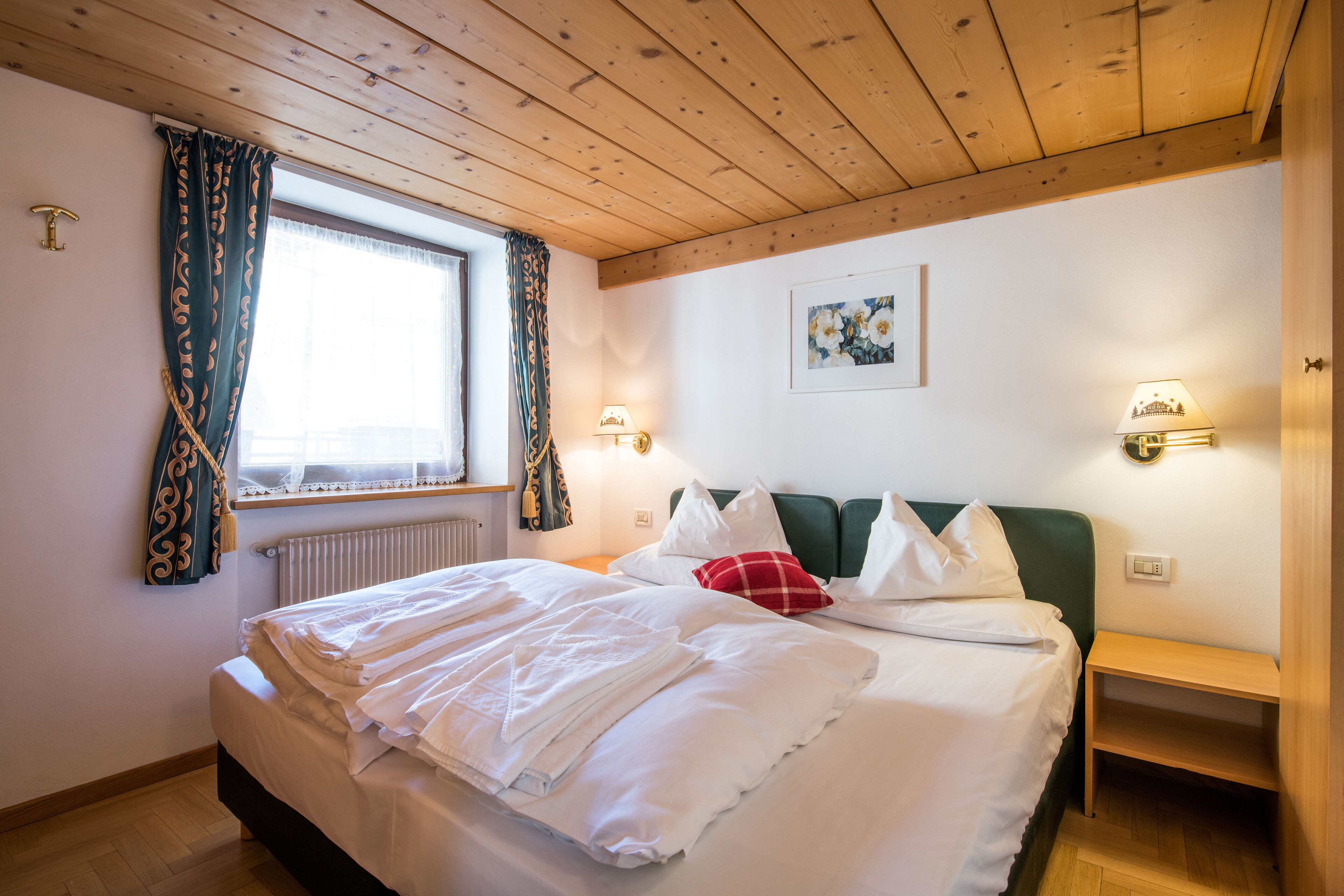 Cama bem arrumada com lençóis brancos, cabeceira verde e almofada vermelha. Há duas luminárias de cabeceira e um criado-mudo de madeira pequena. Uma janela com cortinas é visível, e um quadro emoldurado pendurado na parede.