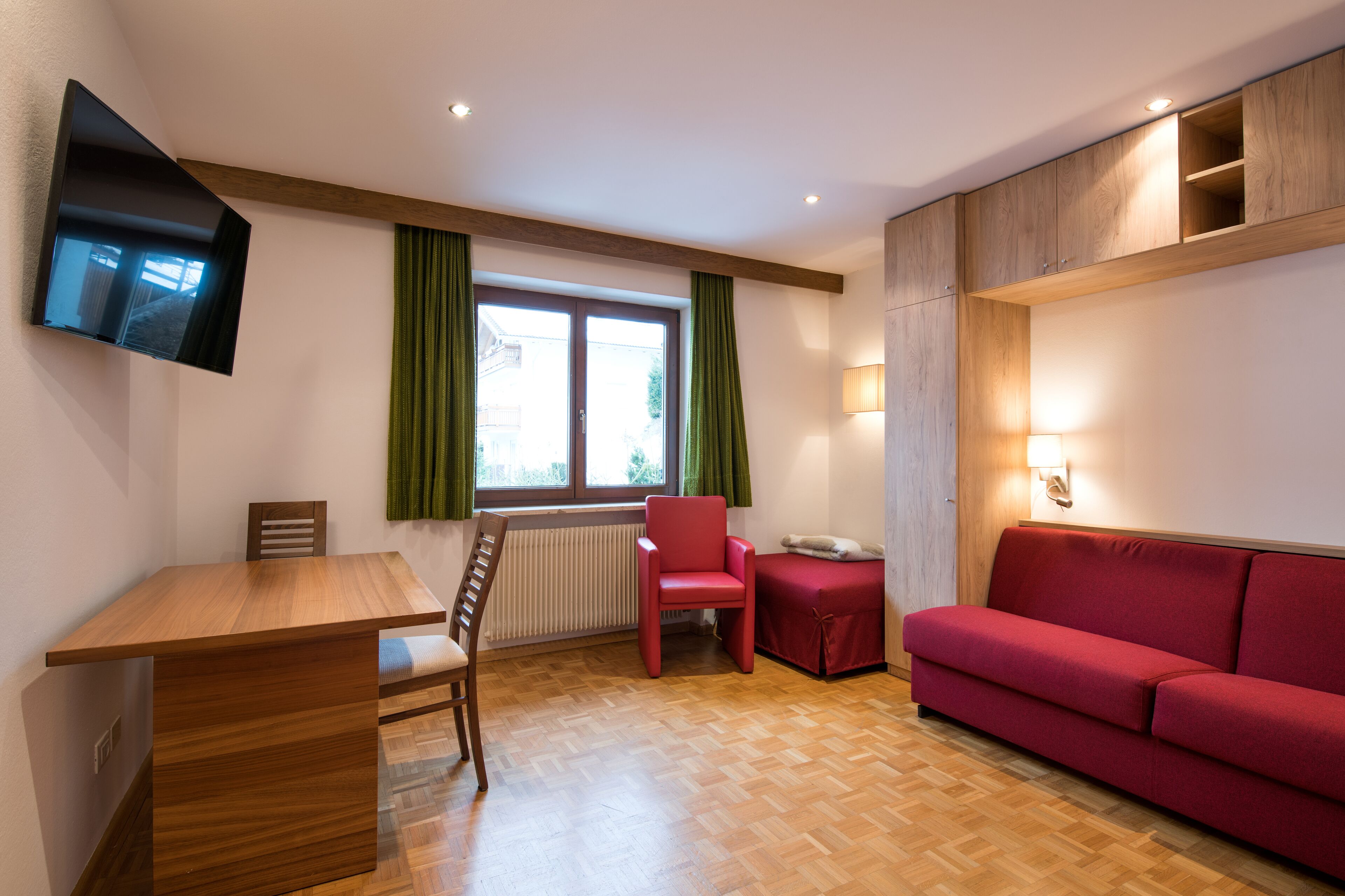 A room with a wooden dining table, a red chair, a red sofa, a TV mounted on the wall, and a window with green curtains.