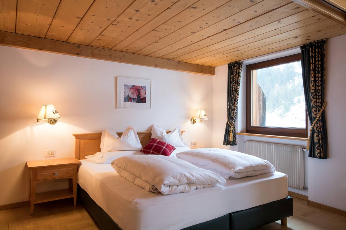 A neatly made bed with white linens and a red pillow, a wooden nightstand with a lamp, and a window with curtains.