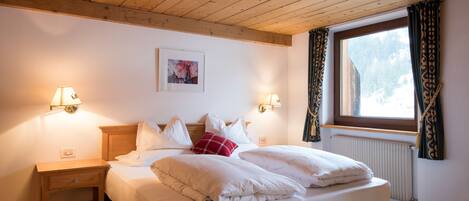 Una cama bien hecha con sábanas blancas y una almohada roja, una mesita de noche de madera con una lámpara, y una ventana con cortinas.