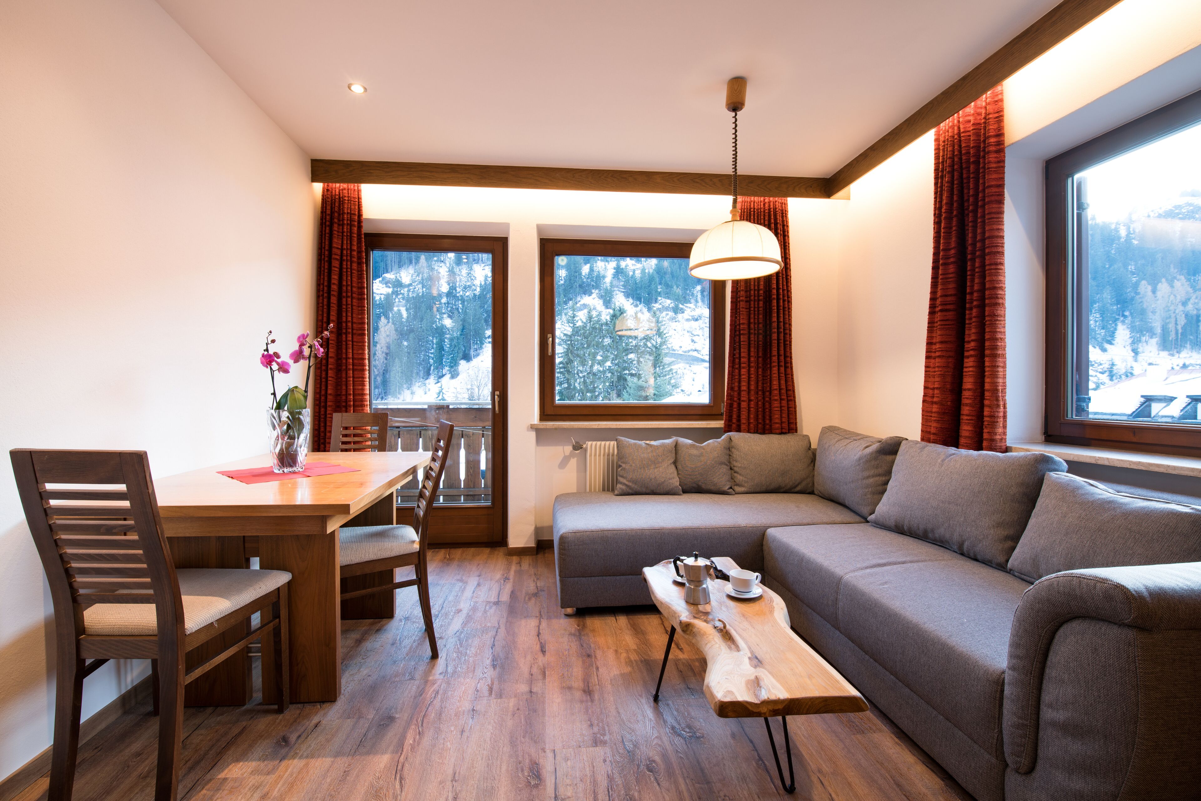 Un salón acogedor con una mesa de comedor de madera, un sofá y vistas a un paisaje montañoso nevado a través de amplios ventanales.