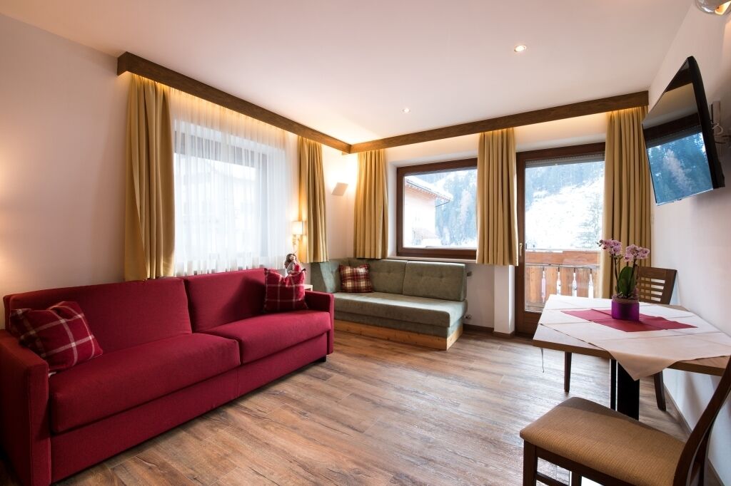 A cozy living room with a red sofa, a green sofa, and a dining table with a chair. There are large windows with curtains, a TV mounted on the wall, and a small table with a potted plant.