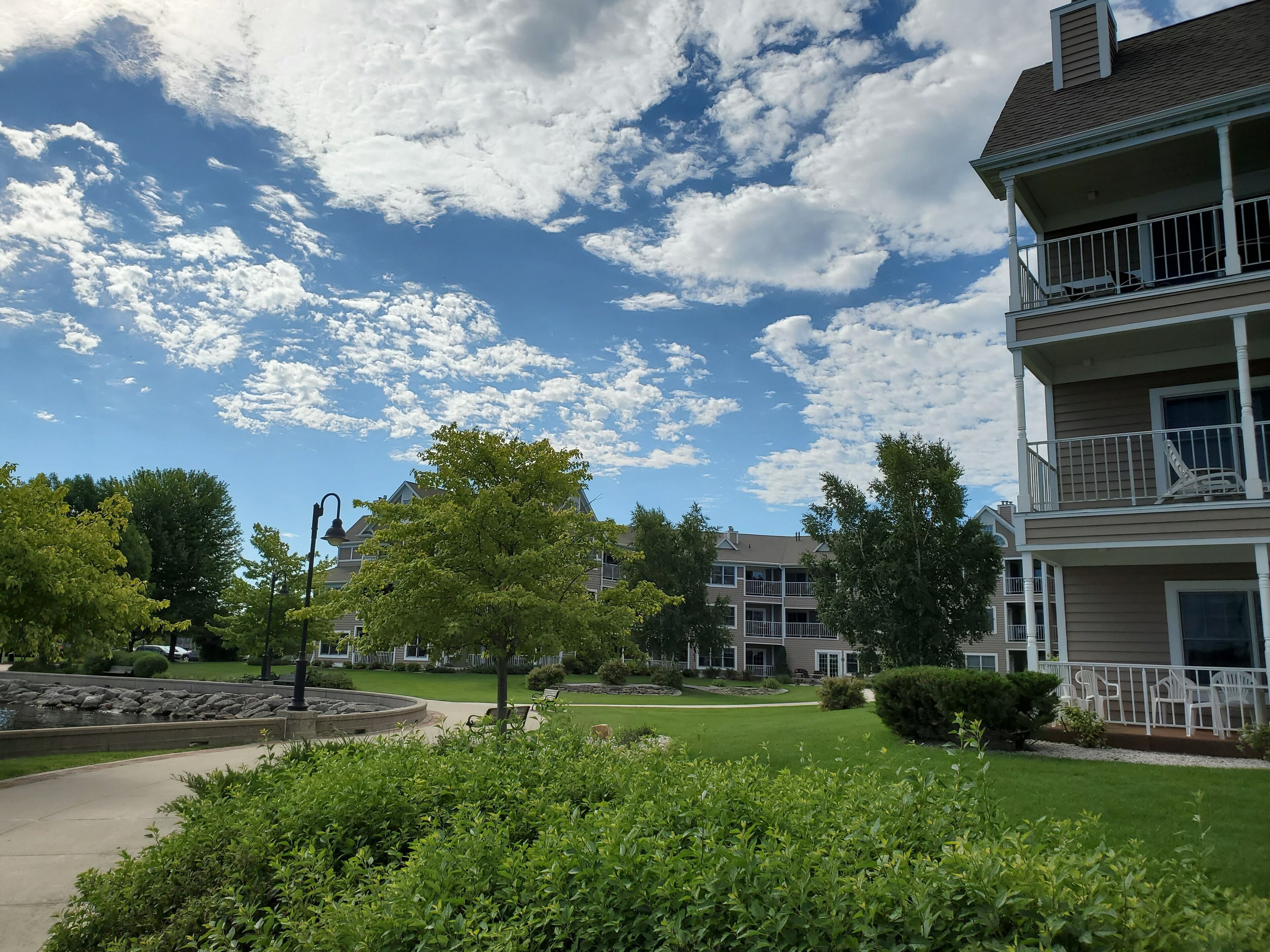 A residential building with a two-story balcony, a landscaped garden with a circular stone feature, and a clear blue sky with scattered clouds.
