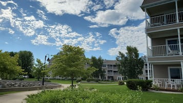 A residential building with a two-story balcony, a landscaped garden with a circular stone feature, and a clear blue sky with scattered clouds.