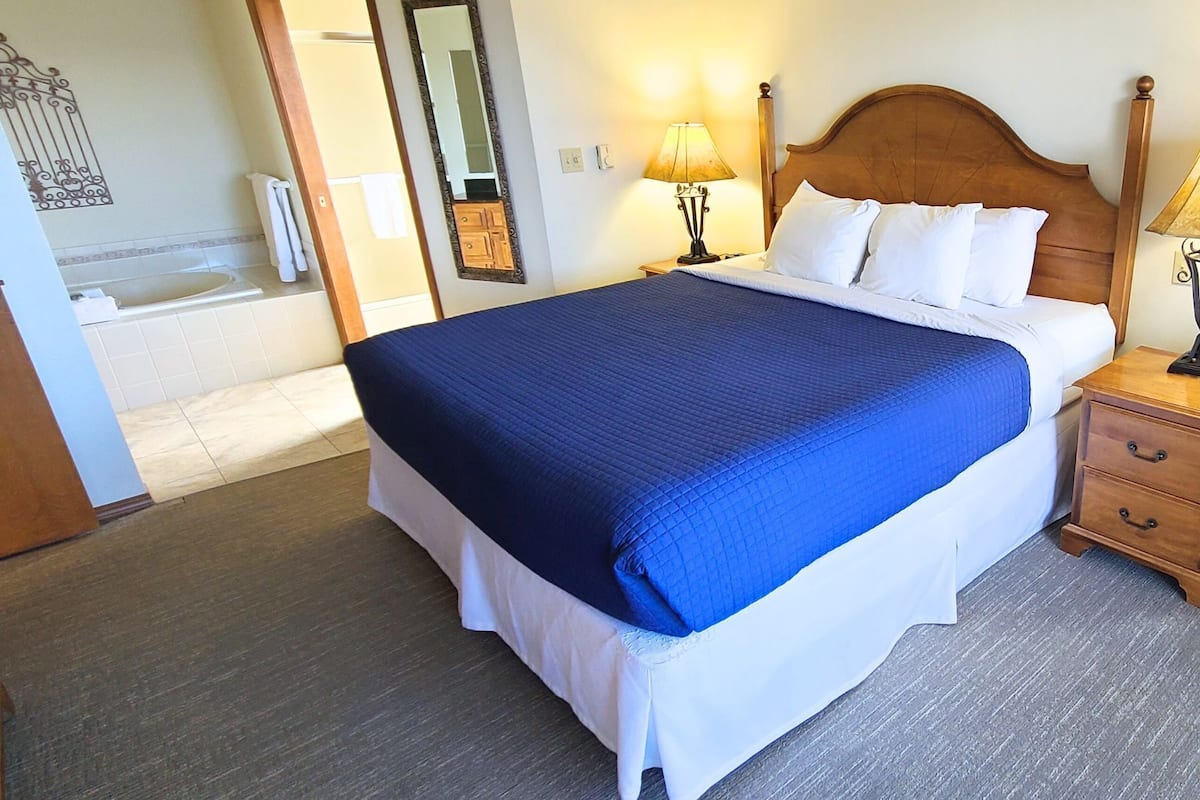 A hotel room with a large bed, a wooden headboard, a nightstand with a lamp, and a bathroom visible through an open door.