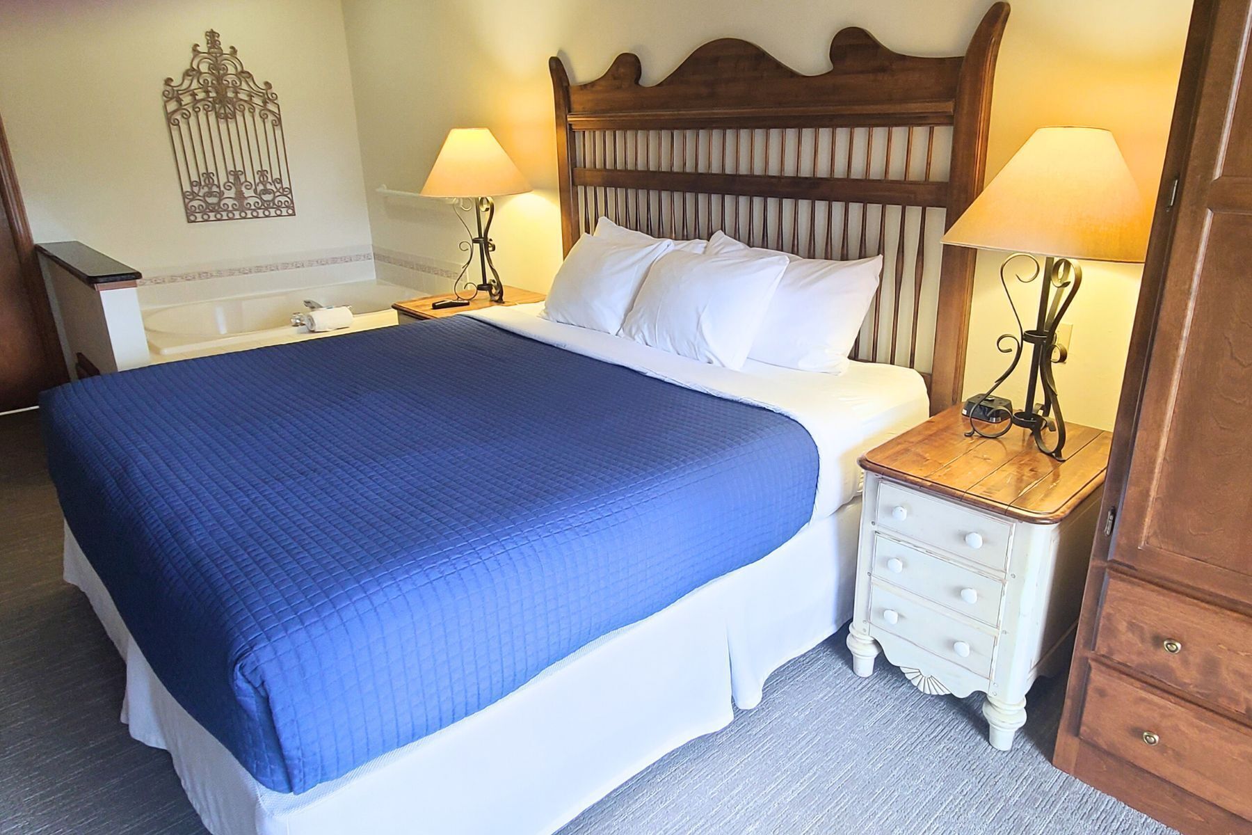 A neatly made bed with a blue blanket, two bedside tables with lamps, a wooden headboard, and a decorative wall piece.