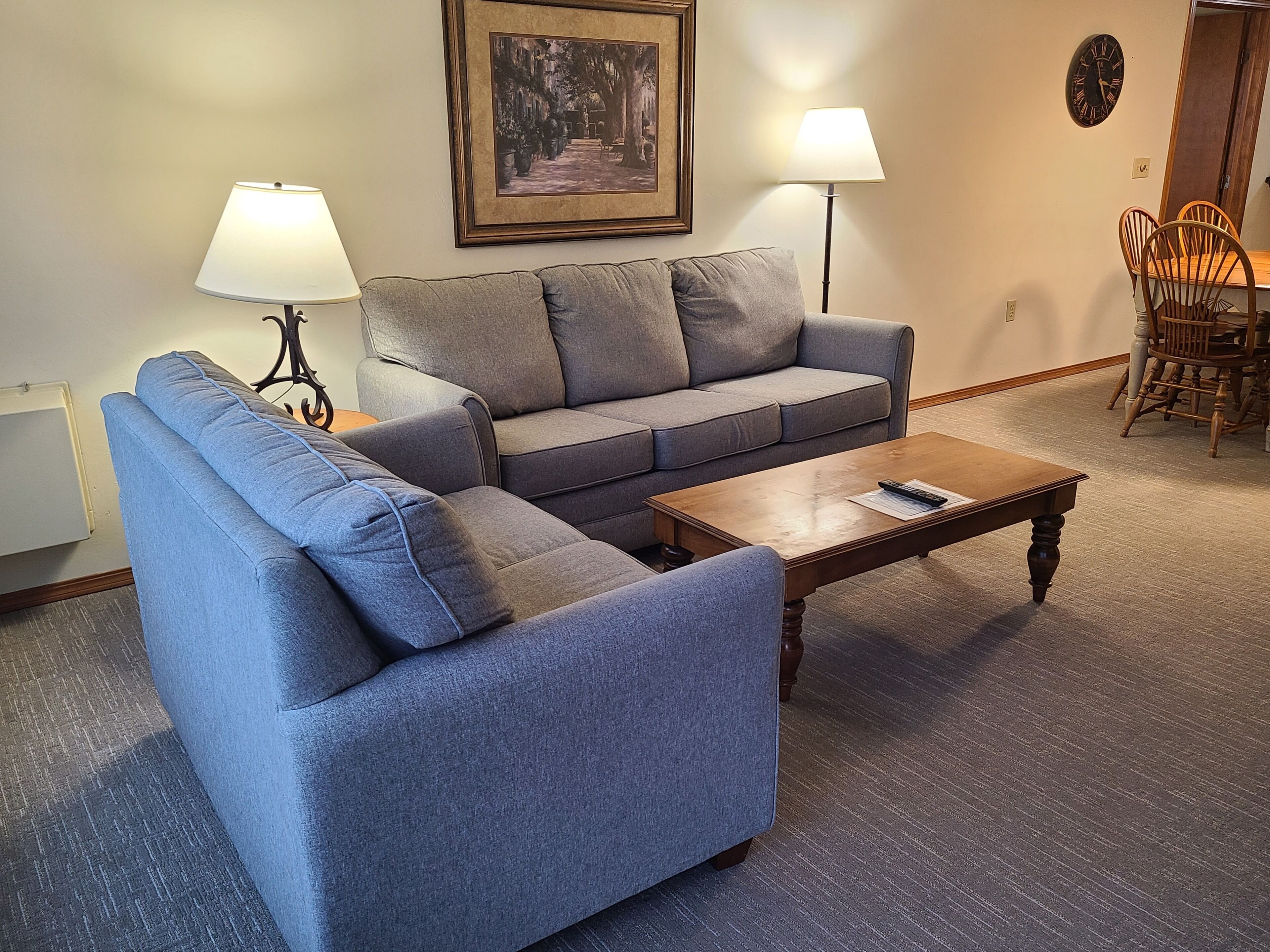 Uma sala de estar com sofá seccional cinza, duas luminárias de chão, uma mesa de centro de madeira e um espaço de jantar com cadeiras e mesa de madeira.