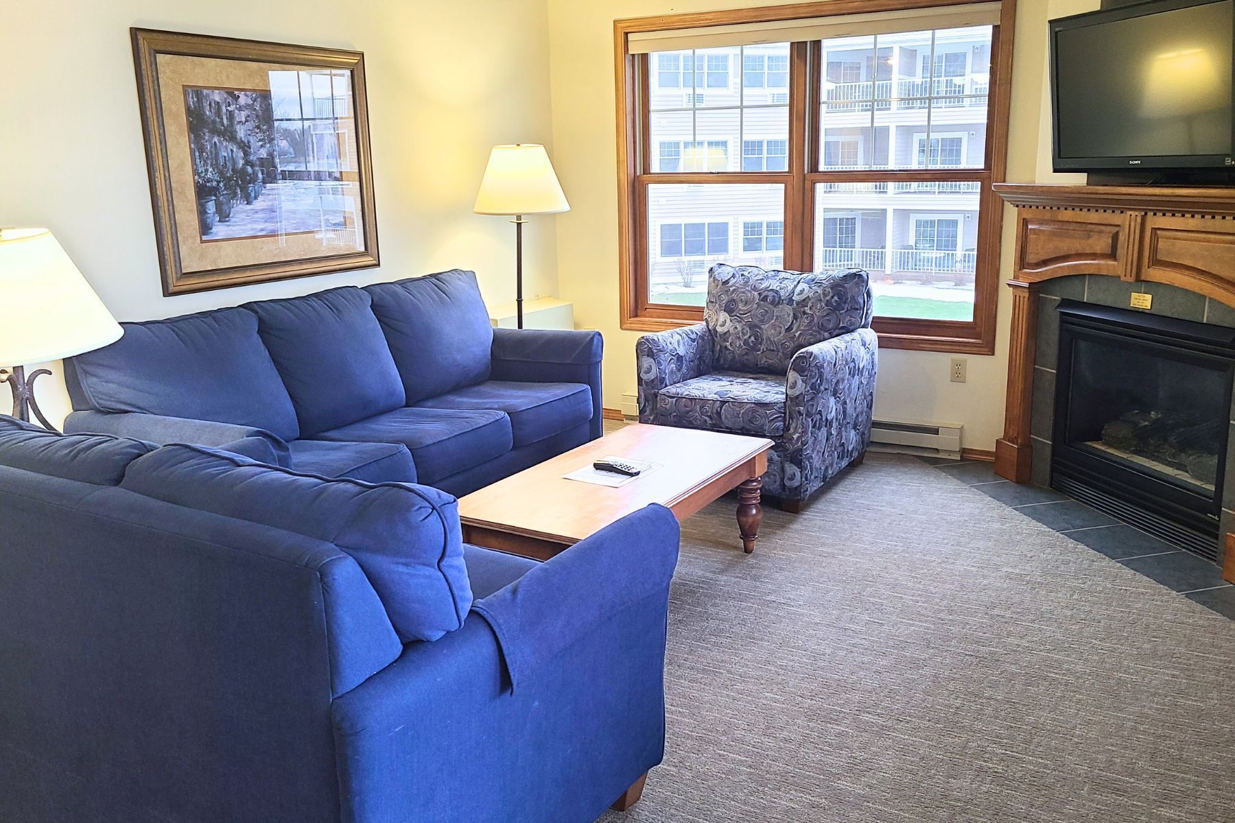 A living room with a blue sofa, a patterned armchair, a coffee table, a fireplace, and a large window with a view of buildings.