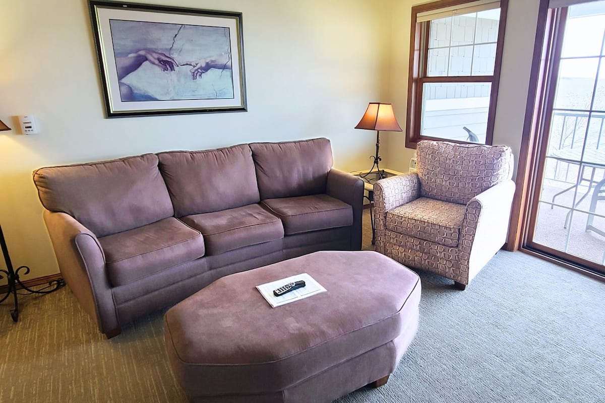 A living room with a brown sofa, a patterned armchair, and a small ottoman. A framed artwork is on the wall, and a lamp is on a side table.