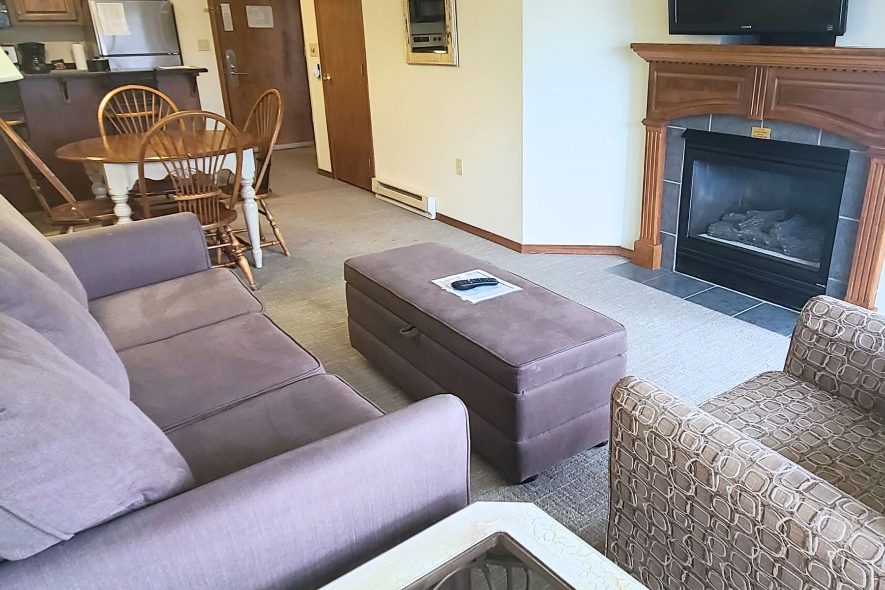 Una sala de estar con sofá, reposapiés, sillón estampado, chimenea y un comedor con sillas y mesa.