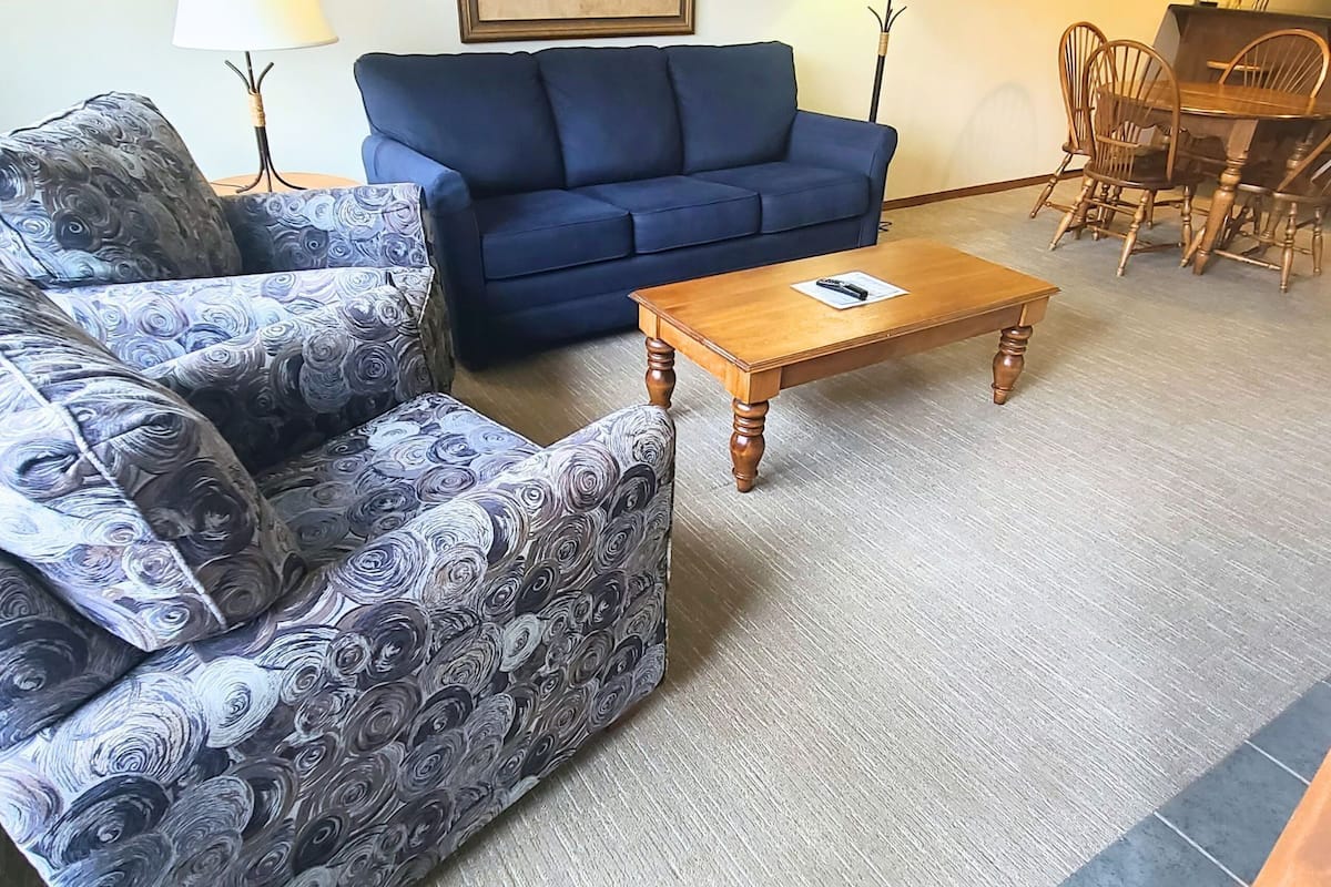 A living room with a blue sofa, patterned armchairs, a wooden coffee table, and a dining area with wooden chairs.