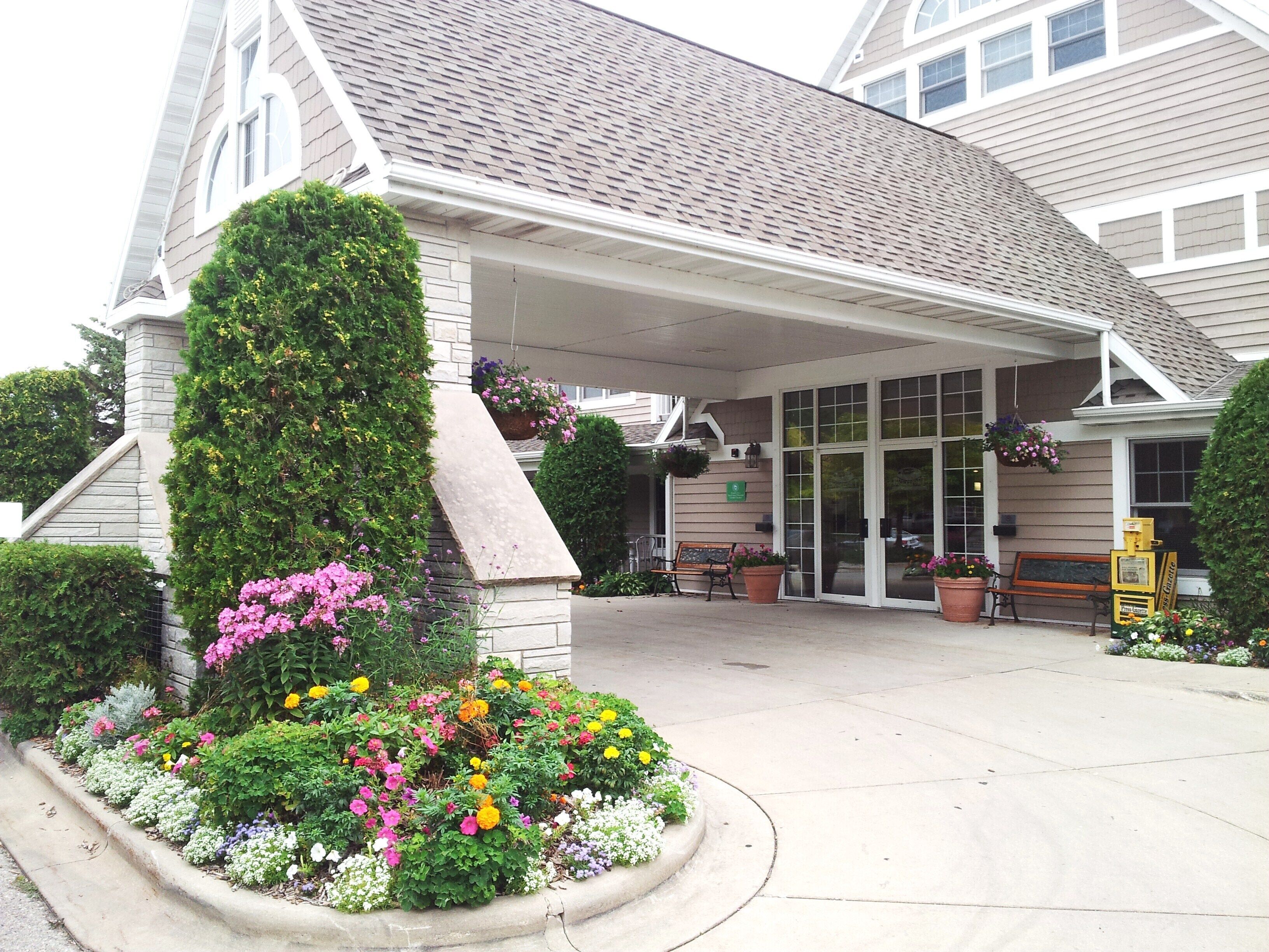 A hotel entrance with a covered porch, a stone chimney, and a variety of colorful flowers in the front garden.