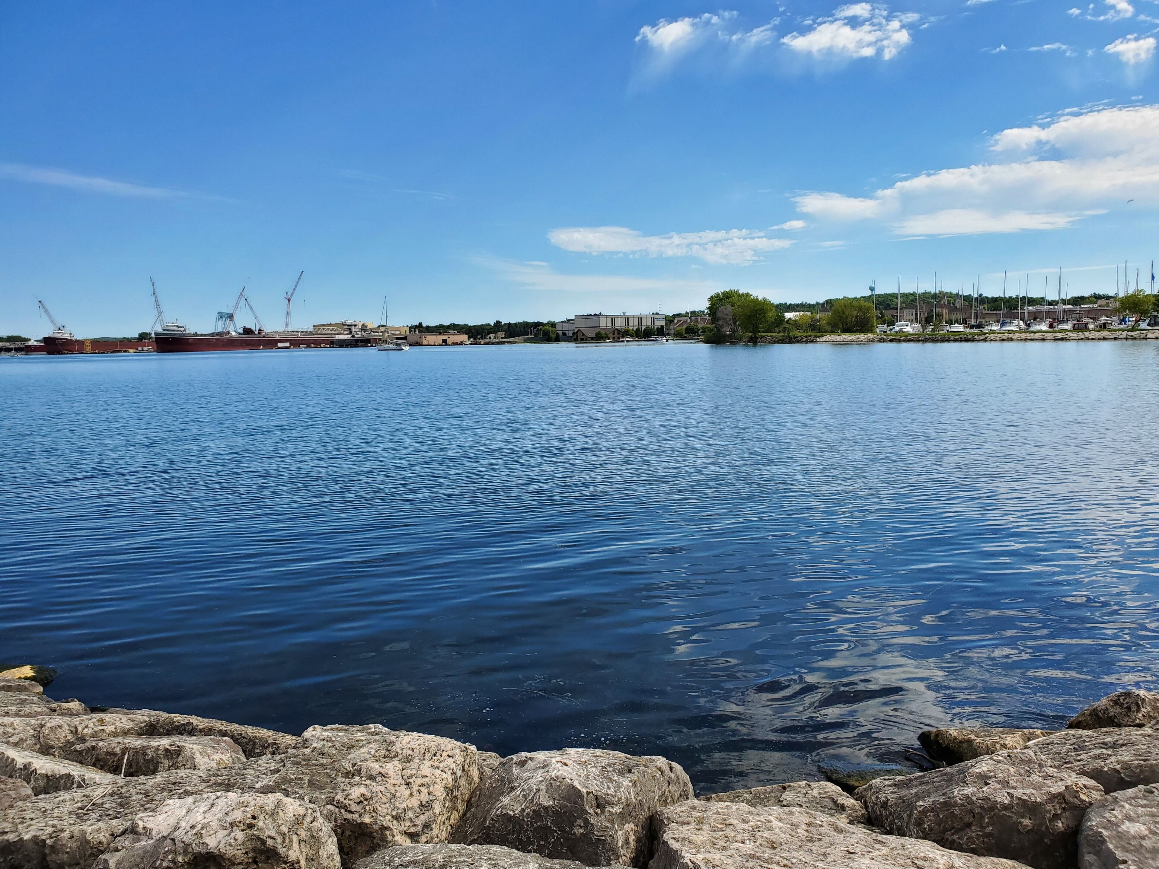A calm waterfront with large rocks in the foreground, a marina with boats and cranes, and buildings along the shore under a clear sky.