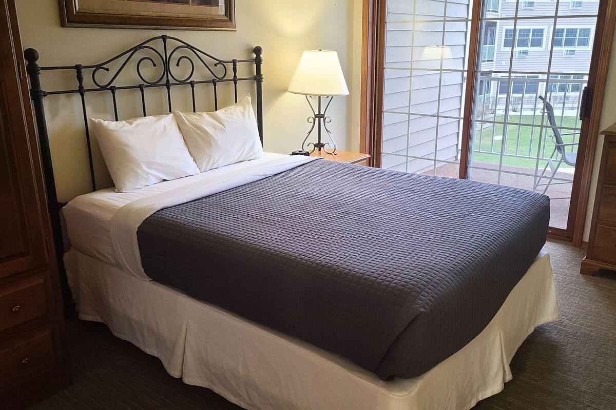 A neatly made bed with white linens and a dark quilted bedspread, a bedside lamp, and a window with a view of a residential area.