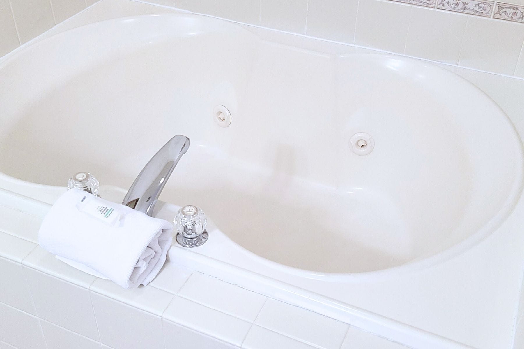 A white bathtub with a silver faucet and two holes, a rolled white towel, and a white tiled wall with a decorative border.