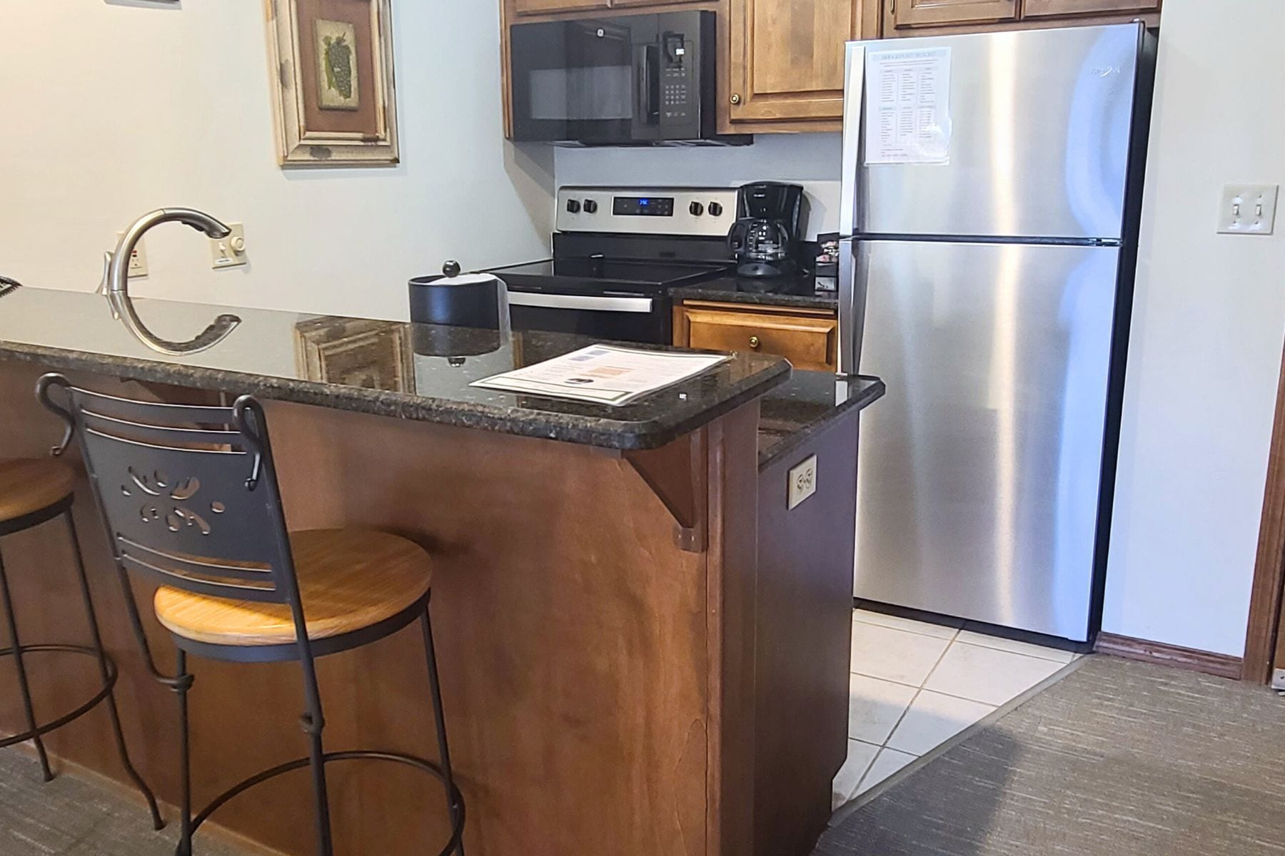 A kitchen with a stainless steel refrigerator, a microwave, a coffee maker, and a sink. There are wooden cabinets and a bar stool with a metal frame.