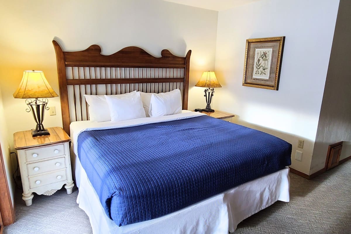 A neatly made bed with a blue quilt, two bedside tables with lamps, a wooden headboard, and a framed picture on the wall.