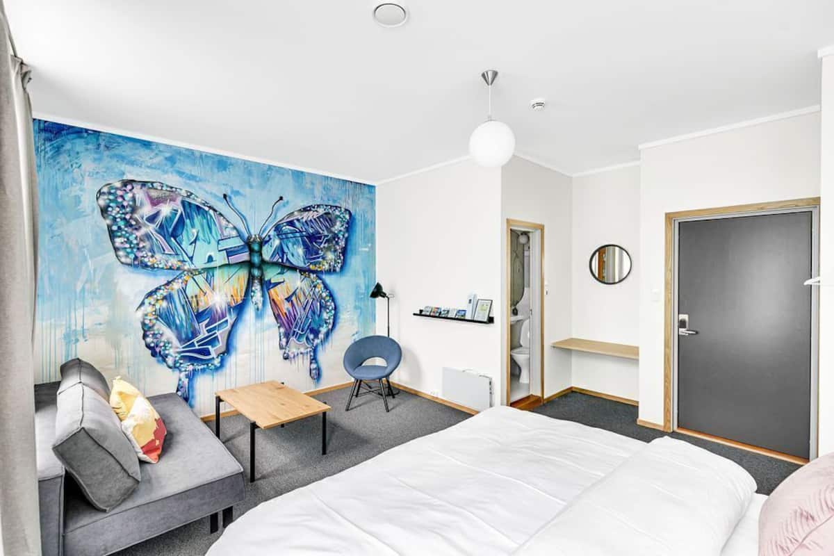 A modern hotel room with a large butterfly mural, a bed, a desk, a chair, a round mirror, and a door.