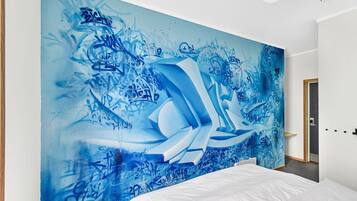 Un dormitorio con un amplio mural abstracto azul en la pared, una cama blanca y una puerta a la derecha.