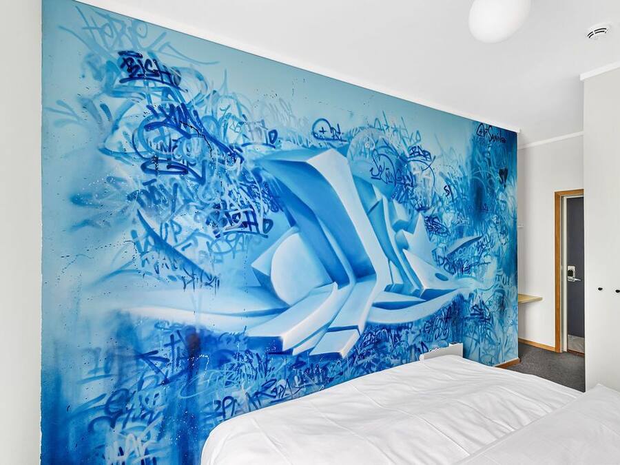 Un dormitorio con un amplio mural abstracto azul en la pared, una cama blanca y una puerta a la derecha.