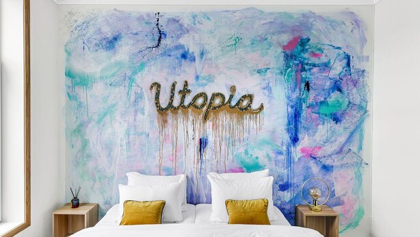 Un dormitorio con un cuadro abstracto grande en la pared, una cama con ropa de cama blanca y almohadas doradas, y dos mesitas de noche con lámparas.