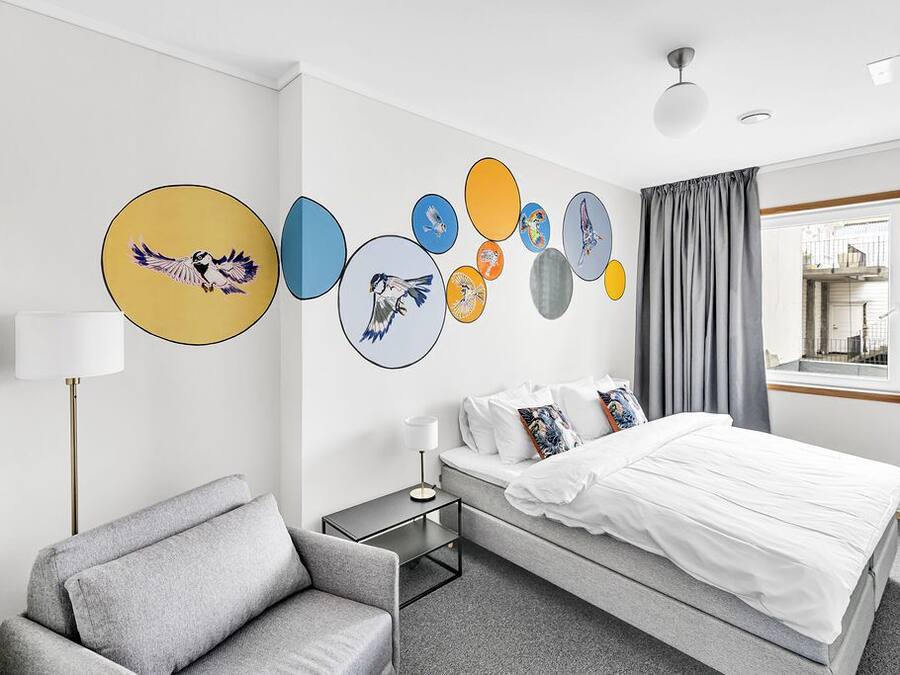 Une chambre d’hôtel moderne avec un lit, un fauteuil gris, une table de chevet et des œuvres d’art murales représentant des oiseaux.