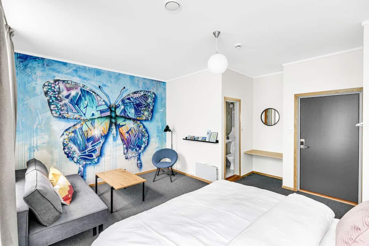 A modern hotel room with a large butterfly mural, a bed, a desk, a chair, and a door.