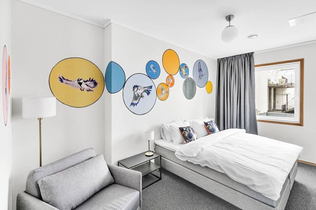 A modern hotel room with a bed, a gray armchair, a nightstand, and wall art.