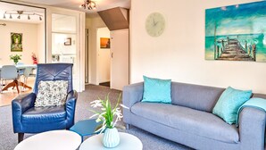 Un salon moderne avec un canapé gris, un fauteuil bleu et une table basse blanche. Un tableau représentant une jetée est accroché au mur, et une horloge est également visible.