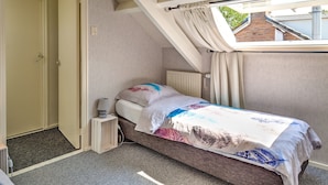 Une petite chambre à coucher, bien rangée, avec un lit simple, une table de chevet, un radiateur et une fenêtre avec des rideaux.