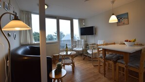 Ein Wohnzimmer mit einer Couch, einem Esstisch mit Stühlen, einem an der Wand befestigten Fernseher und einem großen Fenster mit Vorhängen.
