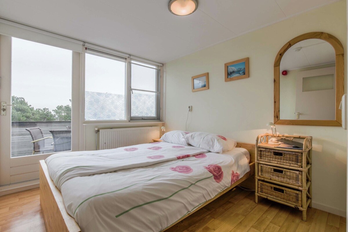 Ein Schlafzimmer mit einem Bett, einem Fenster, einem Spiegel und einem Korbstuhl-Nachttisch.