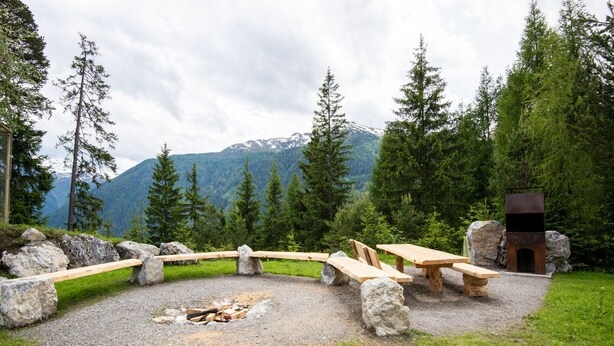 A rustic outdoor seating area with stone benches and a fire pit, surrounded by a natural landscape with mountains in the background.