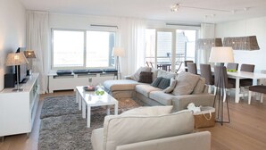 A modern living room with a sofa, coffee table, and dining area. There are large windows, a ceiling fan, and a flat-screen TV. - Modern Apartment in The Hague (The Hague)