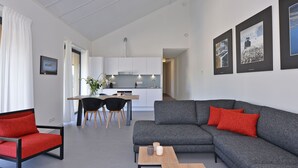 Ein modernes Wohnzimmer mit einem grauen Sofa, roten Kissen und einem Holztisch. Ein Essbereich mit einem weißen Tisch und schwarzen Stühlen ist ebenfalls vorhanden. Im Hintergrund ist eine Küche mit weißen Schränken und einer Edelstahl-Dunstabzugshaube zu sehen.