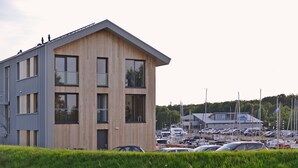 Ein modernes Gebäude mit Holzfassade und großen Fenstern, neben einem Yachthafen mit Booten und Autos.
