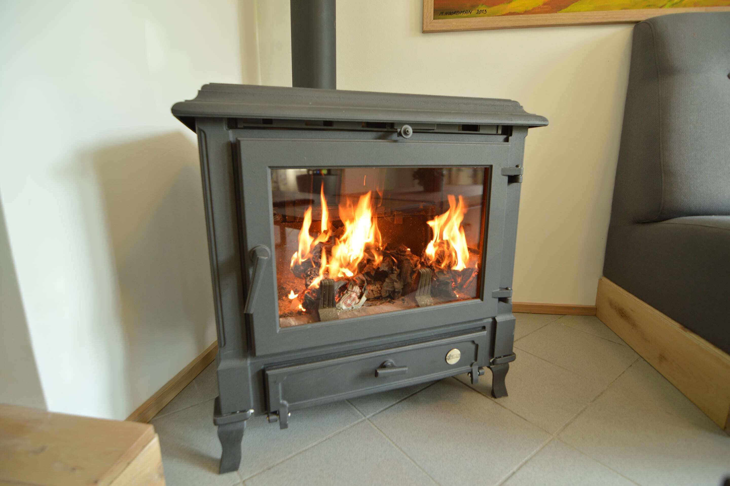 A modern wood-burning stove with a glass front, placed in a corner of a room with a grey sofa and a painting on the wall.